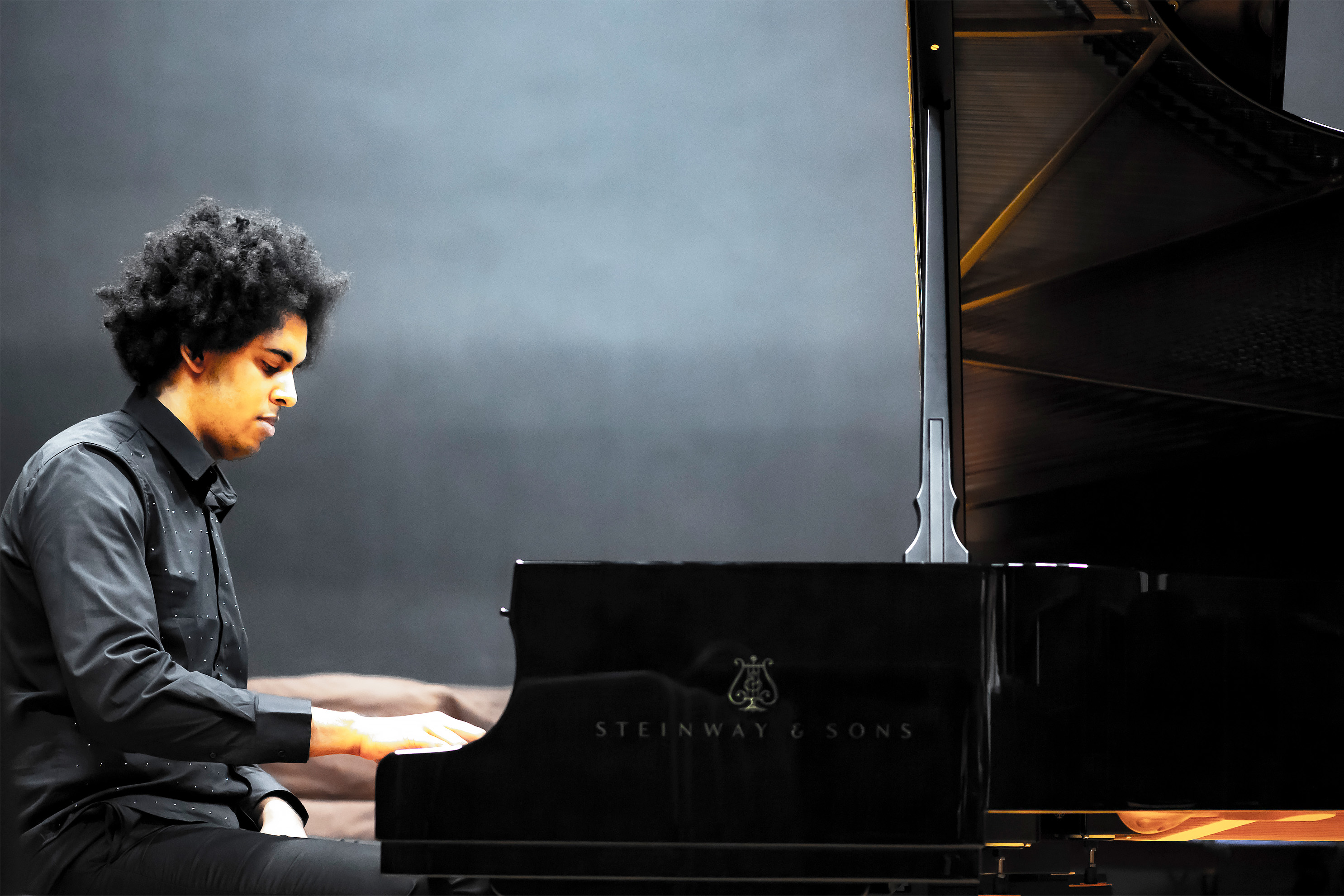 A male student playing the piano, with a dark background behind him.